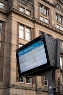 A digital information board displaying bus and tram departure times is positioned in front of a historic building with ornate architectural details and multiple windows.