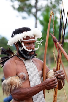 A person is dressed in traditional attire, featuring a headpiece made of white feathers and a necklace. There is white facial paint on their cheeks and ornate beaded decorations on their body. They hold bows and arrows, preparing for possibly a cultural or ceremonial activity.