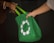 Two people exchanging a green reusable shopping bag with a circular white symbol of birds or hands.