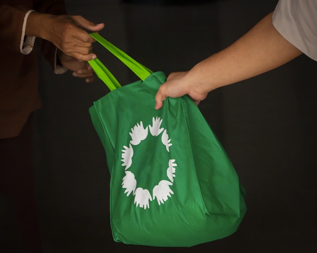 Two people exchanging a green reusable shopping bag with a circular white symbol of birds or hands.