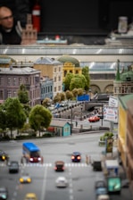 A detailed miniature model of a city street with a bridge, surrounded by various buildings with different architectural styles. There are small vehicles on the road and tiny model trees providing greenery. Signs and traffic lights are present, adding to the urban scene.
