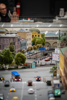 A detailed miniature model of a city street with a bridge, surrounded by various buildings with different architectural styles. There are small vehicles on the road and tiny model trees providing greenery. Signs and traffic lights are present, adding to the urban scene.