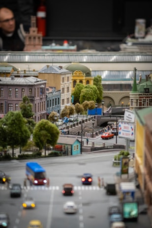 A detailed miniature model of a city street with a bridge, surrounded by various buildings with different architectural styles. There are small vehicles on the road and tiny model trees providing greenery. Signs and traffic lights are present, adding to the urban scene.