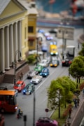 A detailed miniature city scene with a street lined with various small cars, buses, and a motorbike. Trees and a building with columns adorn the sides of the street, while tiny figures of people walk along the sidewalks.