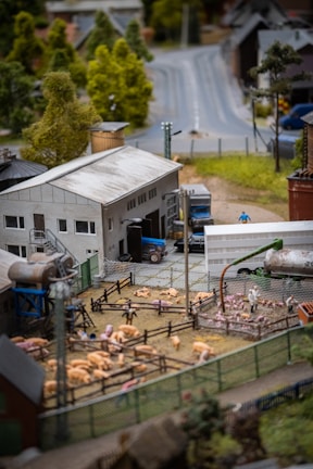 A detailed model of a rural farm scene featuring pigs in fenced enclosures, a grey industrial building, and a truck parked nearby. The setting includes numerous trees, both deciduous and evergreen, along with roads leading into the distance.