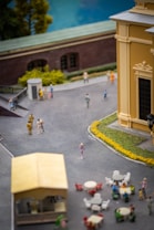 A detailed model scene depicting an outdoor urban setting with miniatures of people walking and interacting. There is a large building with architectural details and a section of greenery along a path. In the foreground, there's a small structure resembling a kiosk or booth and several tables with chairs scattered around.