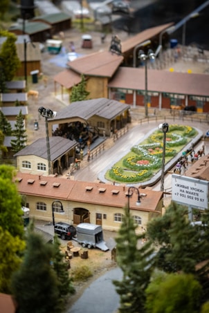 A detailed miniature model depicts a quaint village scene, featuring a small train station with a platform, floral decorations, and a few model vehicles including a car with a trailer. Surrounding the scene are several model trees, adding to the rural atmosphere.