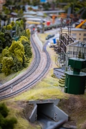 A detailed model railway layout featuring train tracks meandering through a lush, miniature landscape. Scattered buildings, greenery, and hills surround the tracks, creating a vibrant and intricate scene. The focus is on the curve of the tracks as they lead into a small tunnel.
