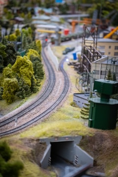 A detailed model railway layout featuring train tracks meandering through a lush, miniature landscape. Scattered buildings, greenery, and hills surround the tracks, creating a vibrant and intricate scene. The focus is on the curve of the tracks as they lead into a small tunnel.
