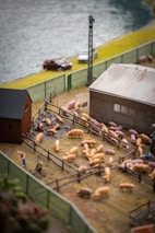 A detailed farm scene features numerous pigs within a fenced area, with a barn and another farm building nearby. The scene includes a small figure tending to the pigs and green fences surrounding the pen. In the background, there's a road with a couple of vehicles next to a river.