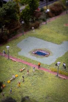 A miniature model of a park scene with small figures engaged in various activities. Several lamp posts line the paths, and a tiny pond is situated in the center. The scene includes people walking and interacting amidst greenery and pathways.