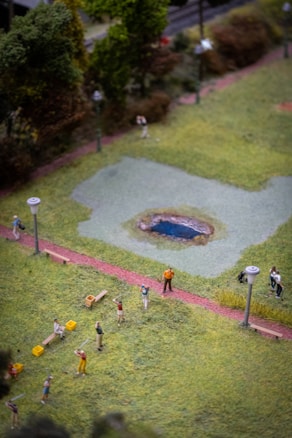 A miniature model of a park scene with small figures engaged in various activities. Several lamp posts line the paths, and a tiny pond is situated in the center. The scene includes people walking and interacting amidst greenery and pathways.