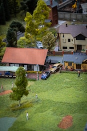A miniature scene depicts a small village with a collection of model buildings, trees, and figurines. In the foreground, there is a grassy area with figures resembling people engaging in activities, possibly playing or walking. Several small buildings with red and gray roofs are present, and a few miniature cars are parked near them. Trees and shrubs are scattered around, giving the setting a peaceful rural appearance.