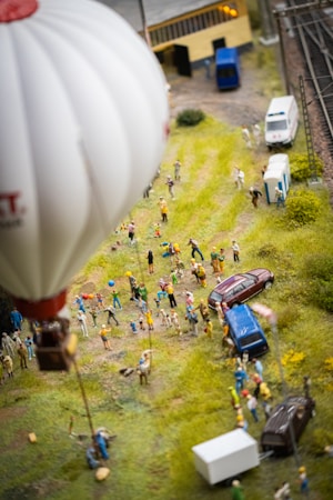A diorama depicting a lively outdoor scene with a hot air balloon in the foreground and numerous miniature figures gathered below it, possibly at a fair or event. There are several vehicles including a blue car, a brown car, and a white van. The setting includes grassy areas, a building, and structures that appear to be part of a railway track on the right.