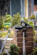 A model scene depicting a damaged building with a burnt, collapsed roof. Two miniature fire trucks with extended ladders are positioned against the building, with small figures of firefighters climbing the ladders. The setting includes surrounding miniature trees and additional model buildings in an urban environment.