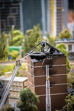A model scene depicting a damaged building with a burnt, collapsed roof. Two miniature fire trucks with extended ladders are positioned against the building, with small figures of firefighters climbing the ladders. The setting includes surrounding miniature trees and additional model buildings in an urban environment.