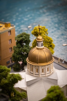 A detailed miniature model of a building with a golden dome and cross on top. Surrounding it are miniature trees and a water body in the background, with a couple of small human figures visible near the water.