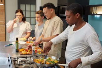 Four people are gathered in a kitchen setting, engaged in cooking and food preparation. One person is stirring a colorful dish in a pan on the stove, while another is chopping vegetables. Two others are observing and chatting, with one sipping from a glass. The countertop is filled with various ingredients and cooking utensils.