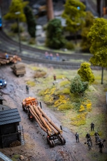 A miniature scene featuring a small truck carrying logs on a dirt road, surrounded by miniature trees and greenery. There are small human figures nearby, some standing and others appearing to interact. The road curves slightly, with a track running parallel, and there are miniature buildings nearby.