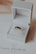 A luxurious ring with dark gemstones is elegantly presented in an open white jewelry box. The box rests on a card with branding and a diamond logo. The background is softly out of focus, enhancing the focus on the ring.