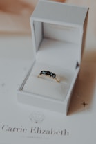 A luxurious ring with dark gemstones is elegantly presented in an open white jewelry box. The box rests on a card with branding and a diamond logo. The background is softly out of focus, enhancing the focus on the ring.