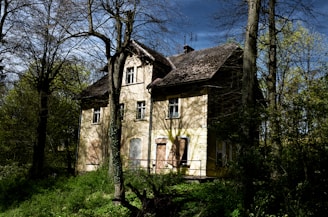 An old, abandoned house is surrounded by dense trees and overgrown vegetation. The building has boarded-up windows and a weathered exterior, suggesting it has been neglected for some time. Sunlight filters through the canopy, casting shadows on the walls.