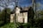 An old, abandoned house is surrounded by dense trees and overgrown vegetation. The building has boarded-up windows and a weathered exterior, suggesting it has been neglected for some time. Sunlight filters through the canopy, casting shadows on the walls.