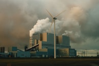 A large industrial facility is emitting thick clouds of white smoke, suggesting active production or power generation. A single wind turbine stands prominently in the foreground, juxtaposing the traditional industrial architecture with renewable energy technology. The sky is overcast with heavy, grey clouds, adding a somber tone to the scene.