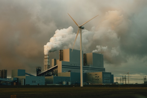 A large industrial facility is emitting thick clouds of white smoke, suggesting active production or power generation. A single wind turbine stands prominently in the foreground, juxtaposing the traditional industrial architecture with renewable energy technology. The sky is overcast with heavy, grey clouds, adding a somber tone to the scene.