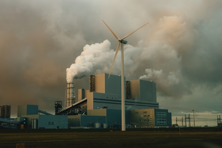 A large industrial facility is emitting thick clouds of white smoke, suggesting active production or power generation. A single wind turbine stands prominently in the foreground, juxtaposing the traditional industrial architecture with renewable energy technology. The sky is overcast with heavy, grey clouds, adding a somber tone to the scene.