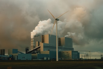 A large industrial facility is emitting thick clouds of white smoke, suggesting active production or power generation. A single wind turbine stands prominently in the foreground, juxtaposing the traditional industrial architecture with renewable energy technology. The sky is overcast with heavy, grey clouds, adding a somber tone to the scene.