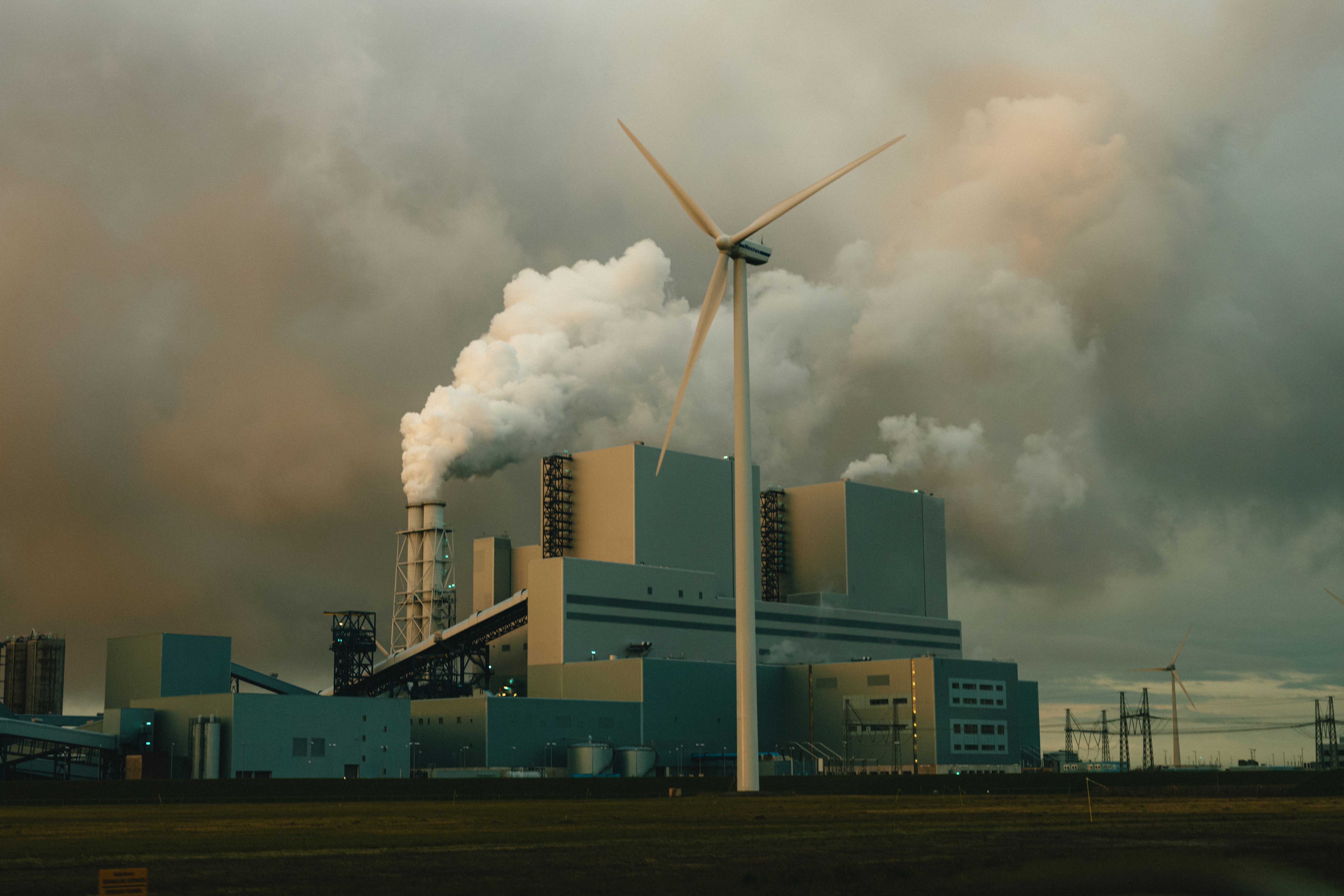 In the heart of Eemshaven's industrial harbor, a lone wind turbine stands tall, asserting its presence beneath the stormy skies of the Netherlands. As the sun dips below the horizon, casting an orange glow, a coal-burning power station looms in the background, with thick smoke billowing into the air.