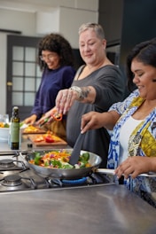 A lively cooking class in progress with diverse participants smiling and chopping fresh vegetables around a large kitchen island.