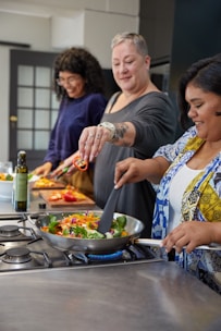 Three people are enjoying a cooking session in a modern kitchen. They are cheerfully preparing a meal, with one person holding a skillet on the stove filled with colorful vegetables. Another person is adding sliced red peppers to the dish, and a third person is cutting more vegetables in the background.
