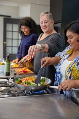 Three people are enjoying a cooking session in a modern kitchen. They are cheerfully preparing a meal, with one person holding a skillet on the stove filled with colorful vegetables. Another person is adding sliced red peppers to the dish, and a third person is cutting more vegetables in the background.