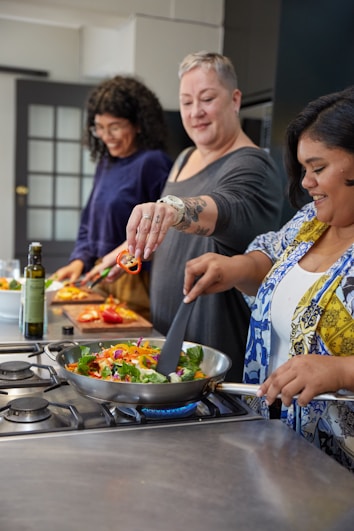 Three people are enjoying a cooking session in a modern kitchen. They are cheerfully preparing a meal, with one person holding a skillet on the stove filled with colorful vegetables. Another person is adding sliced red peppers to the dish, and a third person is cutting more vegetables in the background.