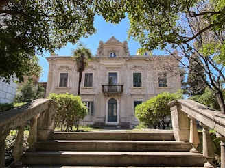 A grand, two-story mansion with ornate architectural details, featuring large windows and a small balcony on the upper level. It is surrounded by lush greenery, including well-maintained bushes and tall trees, with a bright blue sky overhead. Stone stairs lead up to the entrance, framed by decorative railings.