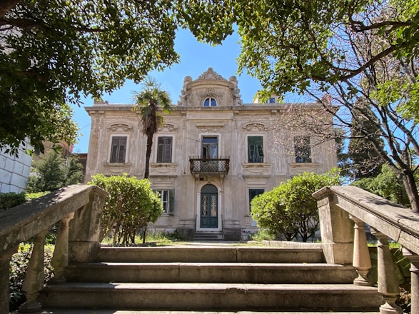 A grand, two-story mansion with ornate architectural details, featuring large windows and a small balcony on the upper level. It is surrounded by lush greenery, including well-maintained bushes and tall trees, with a bright blue sky overhead. Stone stairs lead up to the entrance, framed by decorative railings.