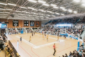 A large indoor basketball arena filled with spectators. A game is underway with players in yellow and dark uniforms competing on a well-lit court. Banners and advertisements are visible along the walls. Bright overhead lights illuminate the scene.
