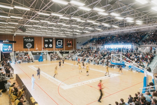 A large indoor basketball arena filled with spectators. A game is underway with players in yellow and dark uniforms competing on a well-lit court. Banners and advertisements are visible along the walls. Bright overhead lights illuminate the scene.