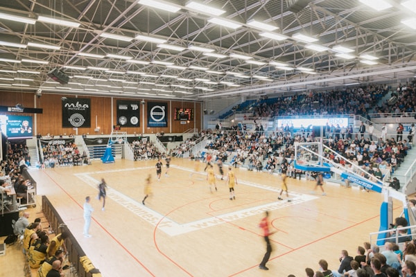 A large indoor basketball arena filled with spectators. A game is underway with players in yellow and dark uniforms competing on a well-lit court. Banners and advertisements are visible along the walls. Bright overhead lights illuminate the scene.