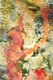 A textured abstract artwork featuring a vibrant mix of colors, including splashes of red, yellow, and green. The composition has a rough, almost organic appearance with visible brushstrokes and a swirling, layered effect.