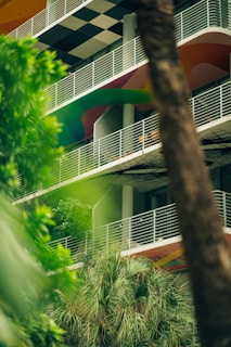 A multistory building with a modern design featuring white railings, colorful curved patterns on the facade, and a black and white checkered ceiling on one level. The foreground contains lush green foliage and palm trees, partially obscuring the view of the building.