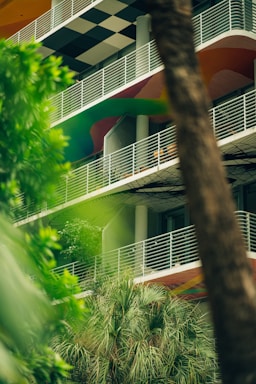 A multistory building with a modern design featuring white railings, colorful curved patterns on the facade, and a black and white checkered ceiling on one level. The foreground contains lush green foliage and palm trees, partially obscuring the view of the building.