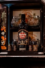 A bottle of Havana Club rum is prominently displayed in the center of a wooden liquor cabinet. Surrounding the bottle are various small bottles of bitters, including flavors like aromatic leaf, coffee, island fruit, and honey. Behind the rum bottle, there are glassware items and a vintage-style Cuban poster with artistic depictions of palm trees and a person. The cabinet includes ornate designs and dark colors, giving it an elegant appearance.