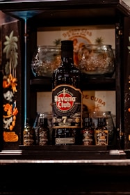 A bottle of Havana Club rum is prominently displayed in the center of a wooden liquor cabinet. Surrounding the bottle are various small bottles of bitters, including flavors like aromatic leaf, coffee, island fruit, and honey. Behind the rum bottle, there are glassware items and a vintage-style Cuban poster with artistic depictions of palm trees and a person. The cabinet includes ornate designs and dark colors, giving it an elegant appearance.