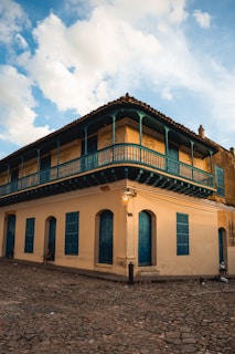 A two-story colonial-style building with blue wooden balconies and shutters, situated on a cobblestone street. The building's walls are painted a warm cream color, complemented by a cloudy sky in the background. A vintage streetlamp is mounted on the corner, near a solitary person sitting by the wall.