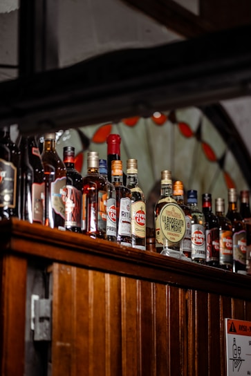 A variety of liquor bottles are displayed on a wooden shelf. The bottles differ in size, shape, and label design, with several brand names visible, including 'Havana Club' and 'La Bodeguita Del Medio'. The background features a decorative stained glass element with red and clear sections.
