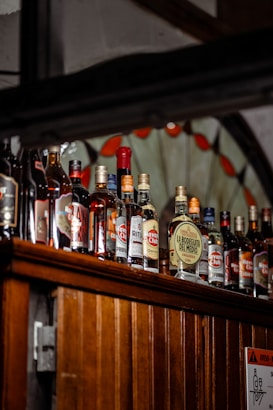 A variety of liquor bottles are displayed on a wooden shelf. The bottles differ in size, shape, and label design, with several brand names visible, including 'Havana Club' and 'La Bodeguita Del Medio'. The background features a decorative stained glass element with red and clear sections.