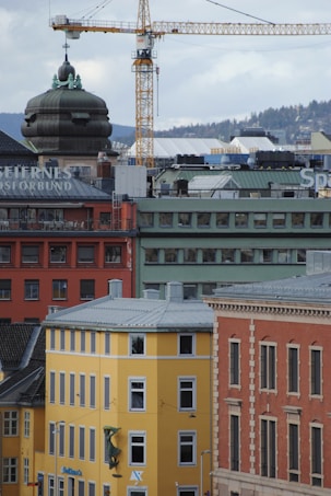 An urban cityscape featuring a mix of colorful buildings, including yellow, red, and green structures. A construction crane towers in the background, indicating ongoing development. The architectural style is varied, with a prominent dome visible on one building. The city is set against a backdrop of hills and a cloudy sky.
